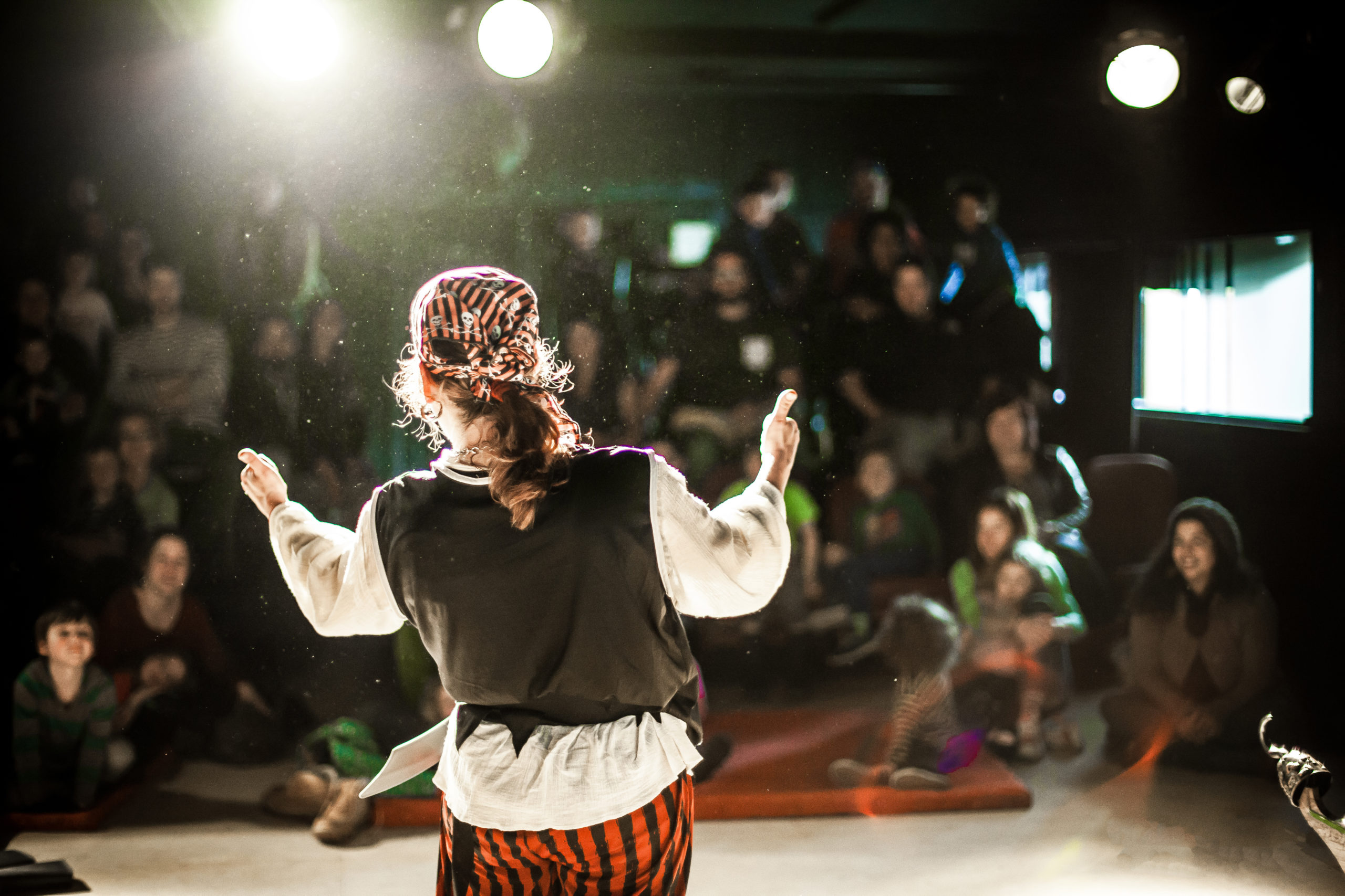 A performing arts entertainer is seen from the back in selective focus, dressed as a pirate on stage during a comedy act with blurry audience at back.